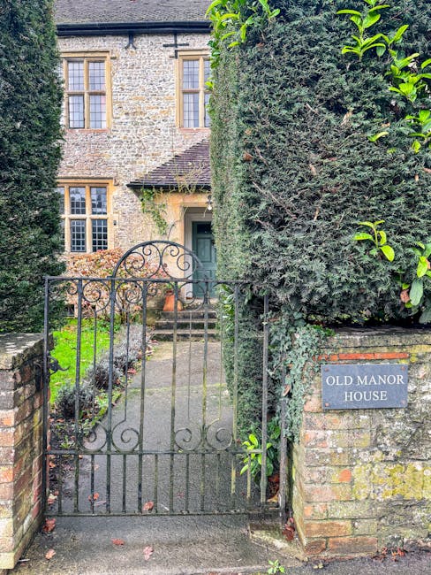 The image shows the entrance to a traditional stone-built house with a black wrought iron gate and fence in the foreground. The gate, which is slightly open, leads to a narrow stone pathway that ascends towards the house's front door. To the right of the gate, there is a brick pillar with a metal plaque engraved with the words 'OLD MANOR HOUSE'. Behind the gate, the property features a well-maintained garden area with small shrubs and plants. The house has multiple windows with white frames, and the stone exterior is partially covered by climbing plants. Tall, dense hedges border the right side of the entrance, providing privacy. The scene is lit by natural daylight, with a mix of greenery and architectural details visible, indicating a quiet residential area suitable for home relocation or furniture transport activities, with [COMPANY_NAME] providing removals services in the area.