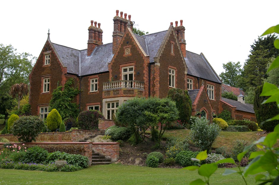 An aerial view of a large, historic brick house with multiple pitched roofs and several chimneys, situated within a green garden with well-maintained lawn and flowerbeds. The house is surrounded by mature trees and pathways, with a paved driveway leading to the front entrance. Adjacent to the property, a removal process is visible where a person wearing a high-visibility vest is carrying a large, wrapped piece of furniture or box towards a nearby van, which is partially visible on the left side of the image. The scene suggests a home relocation or furniture transport activity, with coordination between the mover and the vehicle for efficient loading in a residential setting. The overall environment is well-lit, with natural daylight highlighting the details of the property and the outdoor space, emphasizing the logistics involved in house removals conducted by Man with Van Stamford Hill.