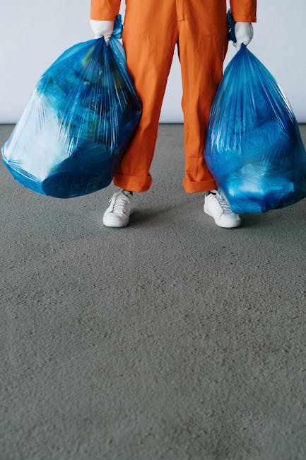 A person dressed in orange work pants and white sneakers is holding two large blue plastic bags filled with various items, standing on a grey carpeted surface near an open doorway. Inside the doorway, a small portion of a room with a light-colored floor and a cardboard box can be seen, indicating a home relocation or moving process. The scene suggests preparation for a house move, with the person likely involved in packing or loading belongings for transport. The background includes a glimpse of a domestic interior, emphasizing the context of furniture transport or removal services. The environment is well-lit, with natural light coming through the open door, highlighting the bags and the person's clothing, which are typical in professional removals and packing tasks, supporting the context of house removals and logistics.