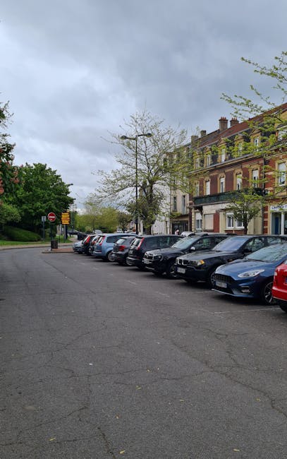 A row of parked cars, including various sedans and hatchbacks, lined up along the pavement outside a residential building with multiple floors and traditional architecture in the Clissold Park area. The sky is overcast with grey clouds, and a few trees with budding leaves are visible, indicating springtime. The scene captures an urban street environment suitable for vehicle loading and unloading activities. As part of house removals or furniture transport, the area appears suitable for parking during home relocation, with visible space for a man with van service such as Man with Van Stamford Hill to facilitate loading or unloading items. No equipment or furniture is visible in this image, but the setting suggests a typical street for moving logistics and packing preparations in the vicinity of the residential properties near Clissold Park.