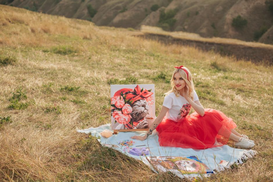 A young woman with blonde hair, wearing a white t-shirt with a floral design, a bright red tulle skirt, and a matching red headband, is seated on a light blue blanket spread out on a grassy hillside.