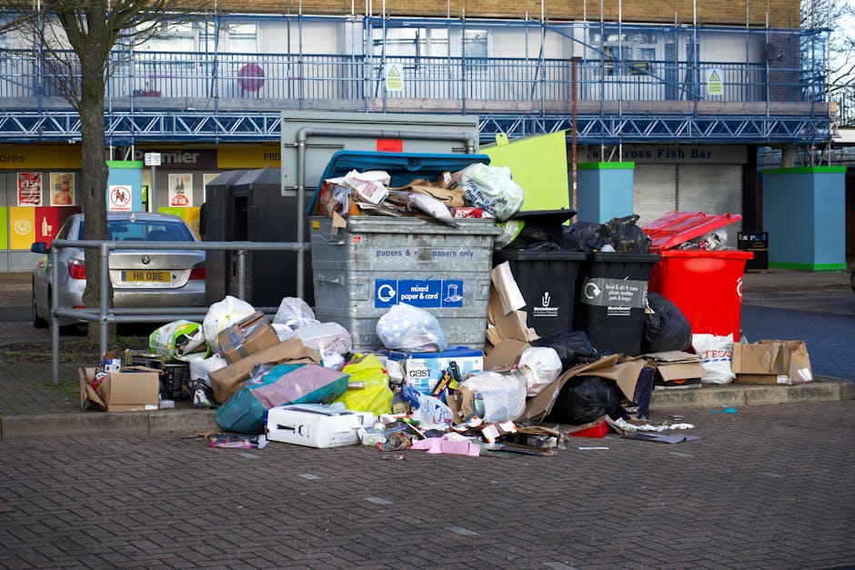 A street scene showing an overflowing public waste collection area with multiple open and closed black, grey, and red wheelie bins, some tipped over, filled with mixed household rubbish including cardboard boxes, plastic bags, packaging materials, and paper waste. There are various cardboard boxes and plastic bags scattered on the pavement around the bins, some containing additional waste. A grey van is parked nearby, partially visible behind the bins, and a metal railing separates the waste area from the sidewalk. In the background, there is a commercial building with large windows and a scaffolding structure, indicating nearby property maintenance or renovation. The scene is well-lit with natural daylight, and the environment appears to be in an urban area. This image reflects the importance of proper waste disposal in home relocation or moving services, as provided by Man with Van Stamford Hill, within the context of controlled waste regulations for residential flats.