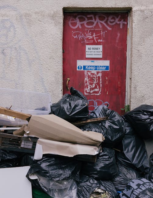 A red metal door with graffiti and multiple signs, including a 'No Money' warning and a 'Keep Clear' notice, is set into a beige wall. In front of the door, there is a pile of black rubbish bags, cardboard boxes, and packaging materials on the pavement, some of which are spilling onto the ground. The scene captures an outdoor area associated with waste disposal near a building, with the trash clearly visible and ready for collection. This image relates to house removals and waste disposal services, illustrating the importance of proper clearance during a home relocation process. Man with Van Stamford Hill, operating within the removals service category, would assist in managing such waste disposal and packing logistics during house moves or estate clearances.