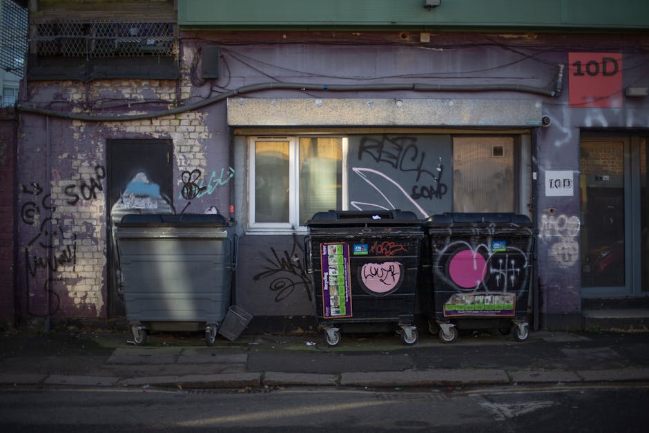 A street scene showing an overflowing public waste collection area with multiple open and closed black, grey, and red wheelie bins, some tipped over, filled with mixed household rubbish including cardboard boxes, plastic bags, packaging materials, and paper waste. There are various cardboard boxes and plastic bags scattered on the pavement around the bins, some containing additional waste. A grey van is parked nearby, partially visible behind the bins, and a metal railing separates the waste area from the sidewalk. In the background, there is a commercial building with large windows and a scaffolding structure, indicating nearby property maintenance or renovation. The scene is well-lit with natural daylight, and the environment appears to be in an urban area. This image reflects the importance of proper waste disposal in home relocation or moving services, as provided by Man with Van Stamford Hill, within the context of controlled waste regulations for residential flats.