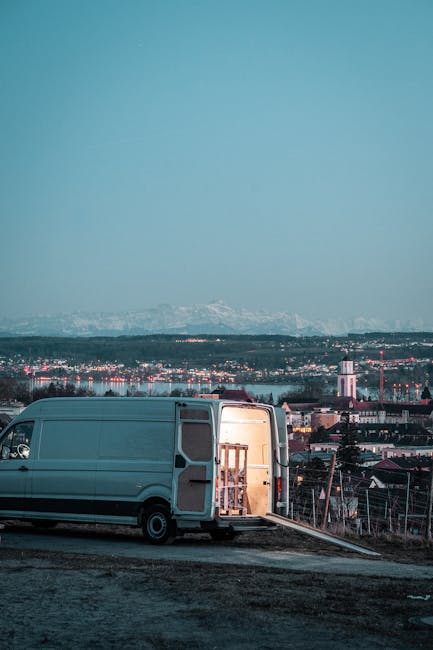 A white panel van with its side door open, parked on a concrete roadway on an elevated area with a cityscape in the background. Inside the van, wooden furniture pieces are visible, possibly wrapped in protective packaging. The van is positioned near a slope leading down to the city, which features residential and industrial buildings, with a tall white tower and a mountain range with snow in the distant background. The scene is illuminated by natural light, suggesting early evening or late afternoon, and the setting indicates a home removal or furniture transport operation related to house moving services. The image supports the context of loading or unloading household items as part of a relocation process, consistent with professional removals by Man with Van Stamford Hill.