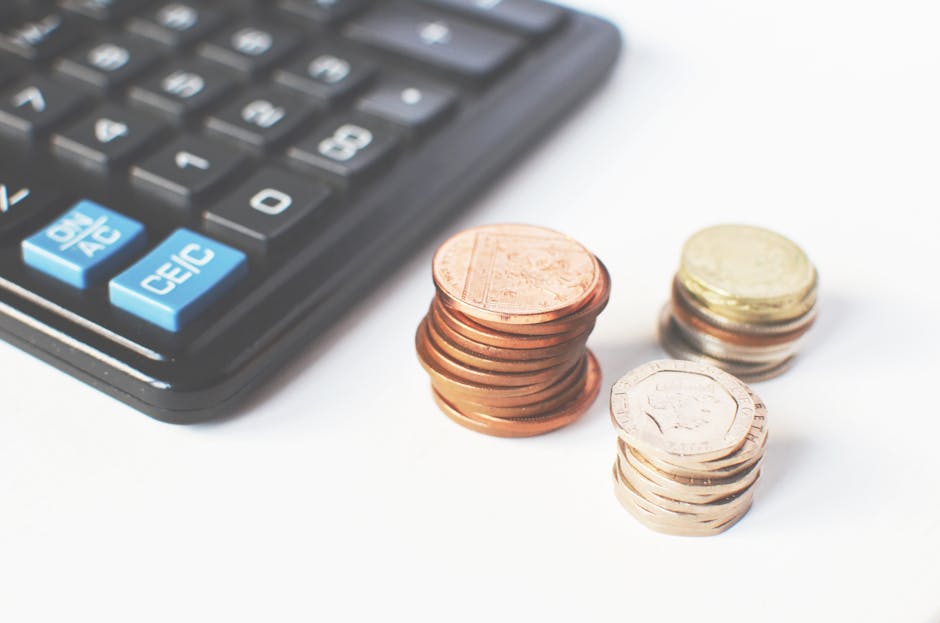 A close-up image showing a black calculator with light blue buttons on its left side, partially out of frame, alongside three stacks of coins in the foreground on a white surface. The first stack contains copper coins, the second stack features a mix of copper and silver coins, and the third stack is composed of silver coins, all positioned to the right of the calculator. The setting suggests an emphasis on financial calculations or budgeting related to house removals or moving costs. The image is relevant to the topic of hidden costs in local quotes for property relocations, as discussed on the page for Man with Van Stamford Hill's removals service, located at https://manwithvanstamfordhill.co.uk.