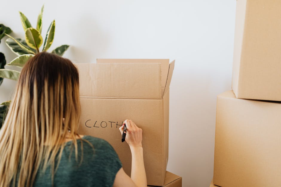A woman with long, straight, light brown hair, wearing a dark green top, is inside a room, writing the word 'CLOTH' on a large cardboard moving box with a black marker. She is positioned with her back to the camera, focusing on labeling the box during the house relocation process. Several other cardboard boxes are stacked nearby, some sealed and others open, indicating packing preparations for a furniture transport or home move. The setting includes a leafy indoor plant to the left, contributing to a tidy, organized environment. The room has white walls and natural lighting, illustrating an indoor packing scene at the start of a house removal service. This image exemplifies packing activities involved in home relocation, with furniture and box preparation supported by Man with Van Stamford Hill, specialists in removals and moving logistics.