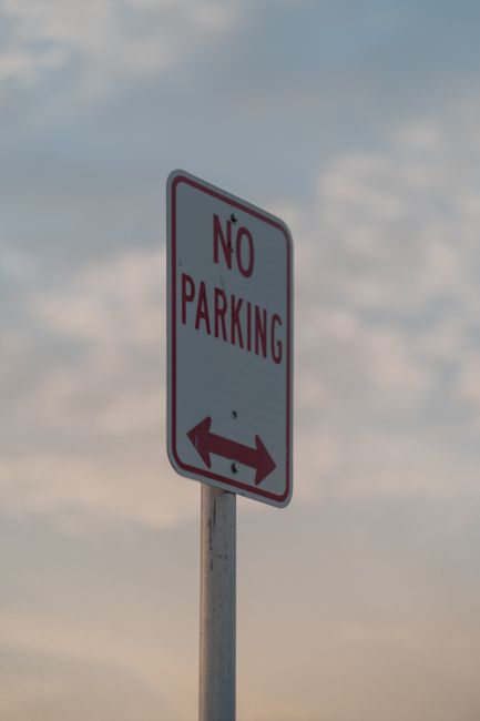 A tall rectangular white 'No Parking' road sign with red borders and red text, mounted on a metal pole situated on a pavement outdoors. The sign indicates parking restrictions and features a double-headed arrow at the bottom pointing left and right, denoting no parking zones on both sides of the sign. In the background, a cloudy sky with patches of blue is visible, suggesting daytime. This image relates to traffic and parking regulations that may impact house removals or relocation services in N16, Stamford Hill, as provided by Man with Van Stamford Hill, with the sign being relevant to parking suspensions and moving logistics in the area.