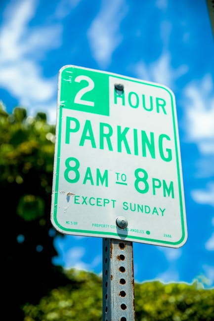 A tall rectangular white 'No Parking' road sign with red borders and red text, mounted on a metal pole situated on a pavement outdoors. The sign indicates parking restrictions and features a double-headed arrow at the bottom pointing left and right, denoting no parking zones on both sides of the sign. In the background, a cloudy sky with patches of blue is visible, suggesting daytime. This image relates to traffic and parking regulations that may impact house removals or relocation services in N16, Stamford Hill, as provided by Man with Van Stamford Hill, with the sign being relevant to parking suspensions and moving logistics in the area.