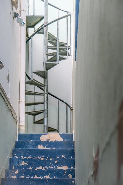 Inside a residential building stairwell with blue-painted steps showing signs of wear and peeling paint, leading up to a screw-in metal spiral staircase with open risers and a black handrail. The stairwell has white walls with some visible damage and piping along the left side, including a showerhead. The scene appears to be part of a home relocation process, where packing materials or furniture are not visible, but the setting suggests preparation for moving tasks. The natural lighting highlights the utilitarian environment, commonly encountered during home removals involving narrow staircases, as managed by companies like Man with Van Stamford Hill.