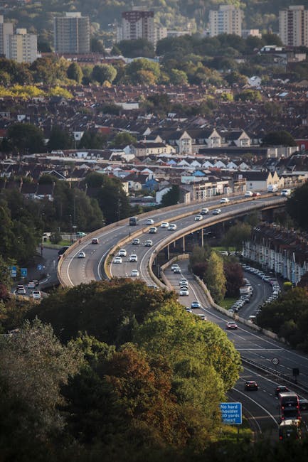 A multi-lane motorway with a gentle curve, shown from an elevated viewpoint surrounded by green trees and foliage in the foreground. Several vehicles, including cars and trucks, are traveling along the road, with some parked on a nearby street in a residential area featuring terraced houses. In the background, there is a densely built urban landscape with numerous buildings, including taller apartment blocks, and a mixture of green spaces and trees. The scene is captured in daylight, with clear weather and natural lighting. This image reflects urban traffic and residential areas, relevant to home relocation and moving logistics as seen in professional removals services by Man with Van Stamford Hill.