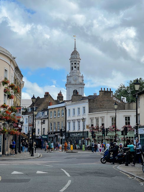 A busy street scene in a town with a historic clock tower and a tall, ornate spire visible in the background under a partly cloudy sky. The street is lined with multi-story buildings, some with decorative facades, balconies, and hanging flower baskets. Pedestrians are walking along the sidewalks, some carrying shopping bags, while others are engaged in conversation or waiting at the bus stop. Several vehicles, including cars, scooters, and a delivery bike, are on the road, while others are parked along the curb. In the foreground, a group of people is loading or unloading items near a van parked at the side of the street, with some objects like boxes and furniture being carried or placed nearby, suggesting a home relocation or moving process. The scene is well-lit by natural daylight, capturing a lively atmosphere typical of an urban shopping and residential area. Man with Van Stamford Hill occasionally conducts furniture transport and packing and moving services, as evidenced by the activity related to loading items into the van.