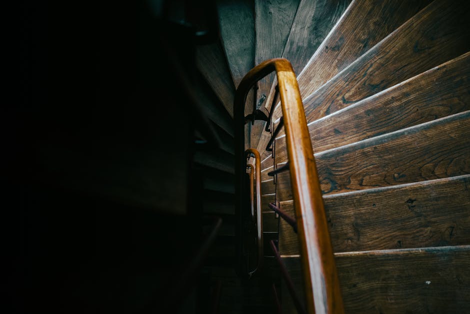 A downward view of a wooden staircase inside a property, showing the staircase’s handrail and steps made of dark, polished wood. The handrail has a smooth, rounded surface and is supported by metal balusters. The stairs ascend into a dimly lit space, with natural light casting soft shadows on the wooden steps. The surrounding walls are dark, emphasizing the rich tones of the wood. This image captures the confined space of a staircase that may pose challenges during a home relocation, illustrating the type of narrow, enclosed environments where careful furniture transport and packing are necessary. Man with Van Stamford Hill offers removals services for such tight staircases, focusing on efficient loading and unloading processes that ensure safe movement of furniture and boxes through limited space areas.