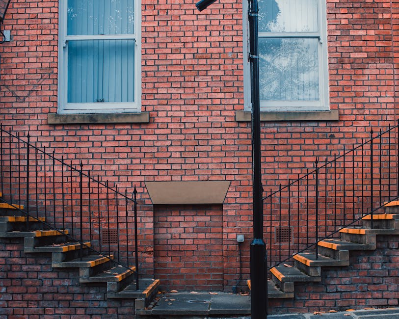 A photograph of a red brick residential building featuring two rectangular sash windows with white frames, positioned above a semi-enclosed area with a small, covered opening set into the brick wall. Outside the building, two exterior staircases with black metal railings and wooden steps, painted with yellow warning strips on the edges, lead up to the first-floor entrances. The staircases are mounted on brick supports and flank a narrow passageway at ground level, which appears to be a recessed doorway or vent. The ground surface is paved with dark concrete, with a few scattered leaves visible at the bottom of the stairs. A black downpipe runs vertically from the roof to the ground between the staircases, attached to the brick wall. Natural daylight illuminates the scene, highlighting the brick textures and the structural components. This setting is part of a home relocation or furniture transport process managed by Man with Van Stamford Hill, aiding with house removals and moving logistics in Stamford Hill.