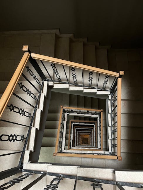 A downward view of a wooden staircase inside a property, showing the staircase’s handrail and steps made of dark, polished wood. The handrail has a smooth, rounded surface and is supported by metal balusters. The stairs ascend into a dimly lit space, with natural light casting soft shadows on the wooden steps. The surrounding walls are dark, emphasizing the rich tones of the wood. This image captures the confined space of a staircase that may pose challenges during a home relocation, illustrating the type of narrow, enclosed environments where careful furniture transport and packing are necessary. Man with Van Stamford Hill offers removals services for such tight staircases, focusing on efficient loading and unloading processes that ensure safe movement of furniture and boxes through limited space areas.